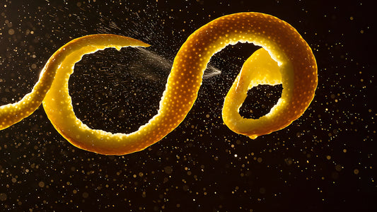 Zeste d'agrume enroulé en spirale sur fond noir, libérant une fine pulvérisation d'huile essentielle dans une lumière rasante, illustration du rapport entre parfum et saison.