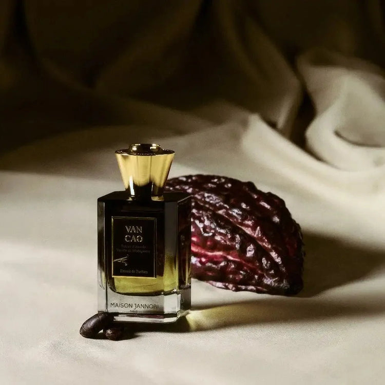 MAISON JANNOBI — the Van Cao bottle resting against a block of stone, surrounded by vanilla pods and a cacao pod, on a dark brown background