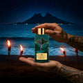 Ahi Pō AB Signature Paris — deux mains tatouées tenant flacon noir cartouche or visible plage volcanique Polynésie nuit étoilée