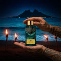 Ahi Pō AB Signature Paris — deux mains tatouées tenant flacon noir cartouche or visible plage volcanique Polynésie nuit étoilée